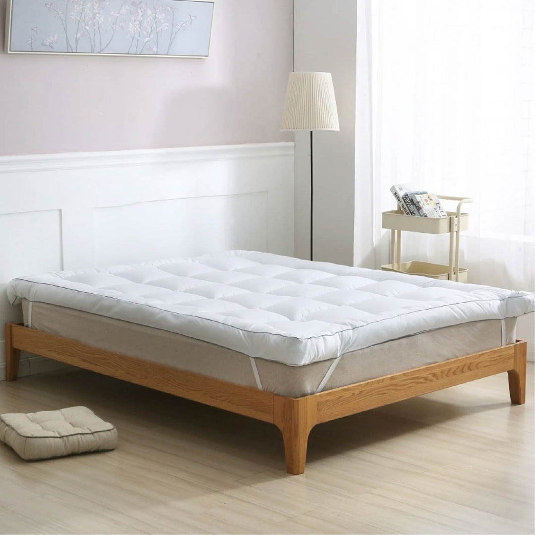 White quilted mattress topper with square stitching placed on a wooden bed frame in a bright bedroom setting.